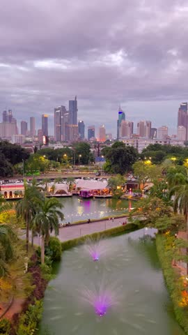 A stunning evening view of the Jakarta city skyline, featuring a tranquil lake with illuminated fountains and lush greenery. The scene captures the transition into twilight as the city lights begin to glow under a purple-hued sky.