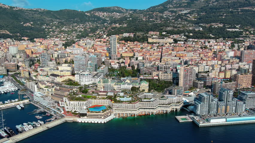 4K aerial view of Monaco vertical cityscape with Casino Square, Port Hercule, rainbow rooftop heliport, dense high-rise towers climbing the mountainside above the Mediterranean coast.