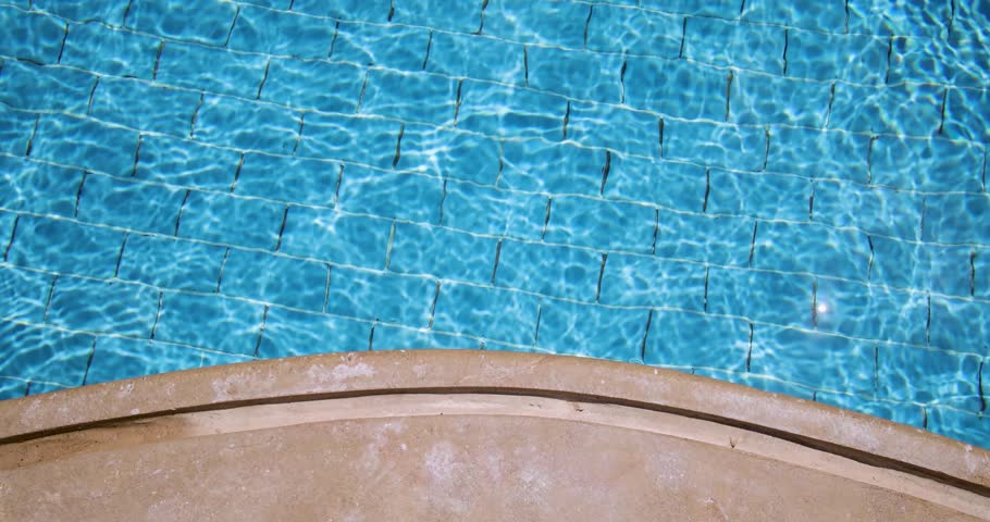 Slow motion footage of shimmering blue swimming pool water with gentle ripples reflecting sunlight across tiled flooring beside a curved pool edge at a luxury resort creating a peaceful relaxing 