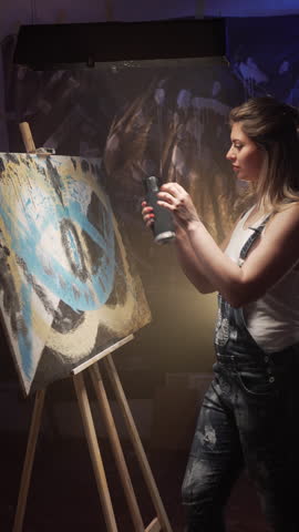 Female artist is painting picture with paint spray can spraying on canvas in her dark modern studio.