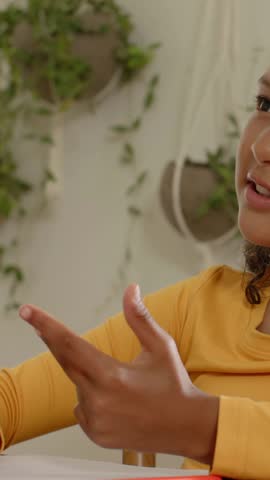 Vertical video: Leaning child in yellow gesturing at table with paper donning headset listening. Girl, notebook, headphones, gesture, conversation, study, vertical video