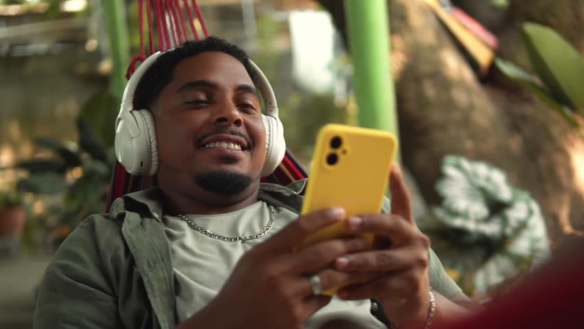 Young man wearing headphones enjoying music and using his smartphone while resting on a hammock outdoors