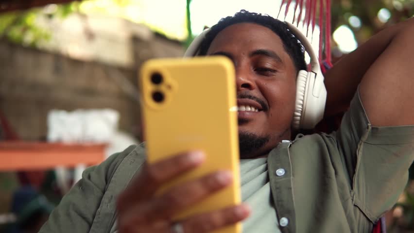Young man wearing headphones relaxing on a hammock and using his smartphone during a sunny day