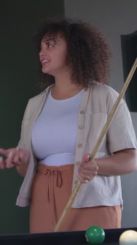 Vertical video: Adjusting cue, woman in white tank beige shirt aiming to sink 3 ball at pool table. Billiards, stick, focus, recreation, home, ambient, vertical video