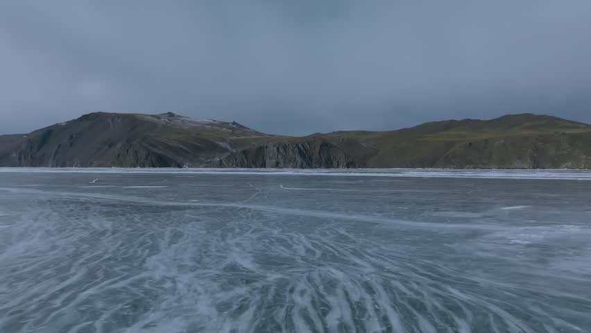 Wide winter landscape footage of cracked blue ice on frozen Lake Baikal with rocky mountain shoreline and overcast sky, natural background in Siberia, Russia