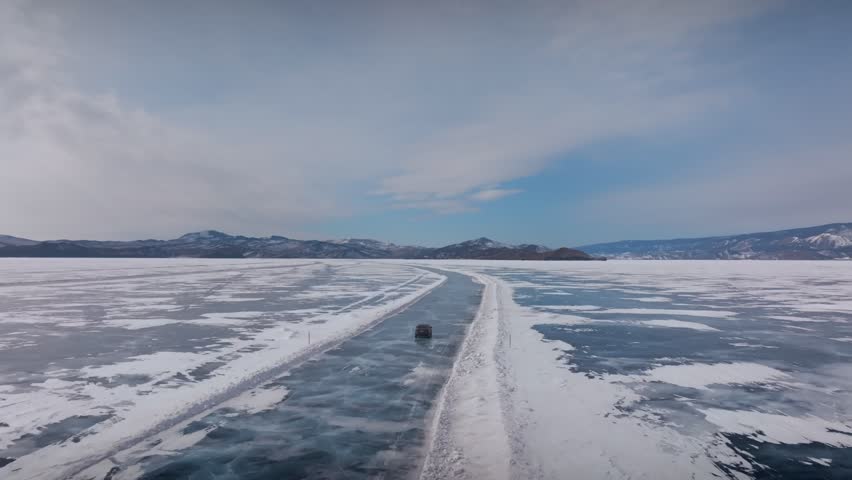 Drone footage of a vehicle driving along a cleared ice road on frozen Lake Baikal in winter, with snowy shores, mountains on horizon, and blue sky above