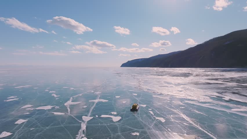 Aerial winter footage of a hovercraft traveling across the frozen ice of Lake Baikal with natural cracks and snow patterns, mountains on the shore, Russia