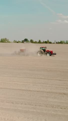 Vertical drone shot of a modern tractor cultivating soil and sowing seeds on a large farm. Clouds of dust trail behind the agricultural machinery under a clear blue sky in springtime