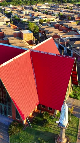Vertical aerial view of Catholic church with red roof in residential neighborhood. Religious building surrounded by houses, streets and greenery under warm golden hour sunlight.