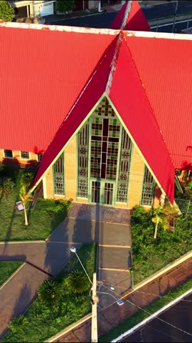 Vertical aerial view of Catholic church with red roof and Virgin Mary statue in residential neighborhood. Religious building surrounded by streets, greenery and urban houses under warm sunlight.