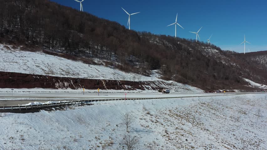 Wind turbines spin atop a snowy mountain ridge above a highway with traffic in this rightward aerial tracking shot of a West Virginia winter landscape under a bright blue sky.
