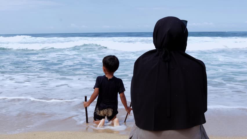 Wide shot of a modest Muslim woman sitting on the sand, looking at her child playing near the ocean waves.