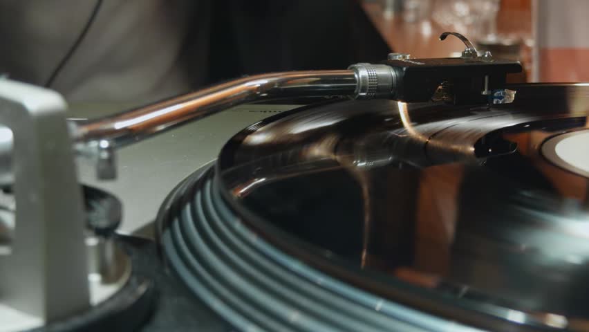 close-up of a vinyl record player with a rotating vinyl record, a tonearm with a pickup needle, DJ hands changing the record, remote control, bar