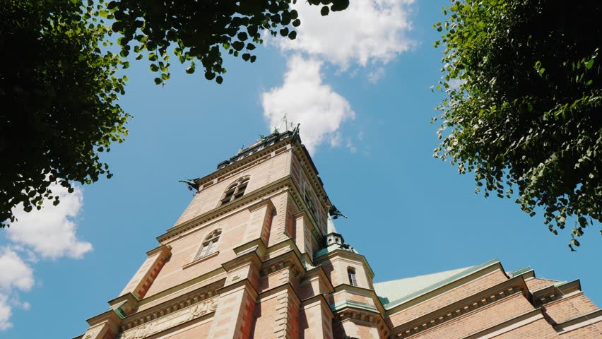Dramatic low angle shot of historical gothic cathedral architecture. Dynamic camera movement reveals red brick tower and decorative spires framed by lush green leaves under bright summer clouds.