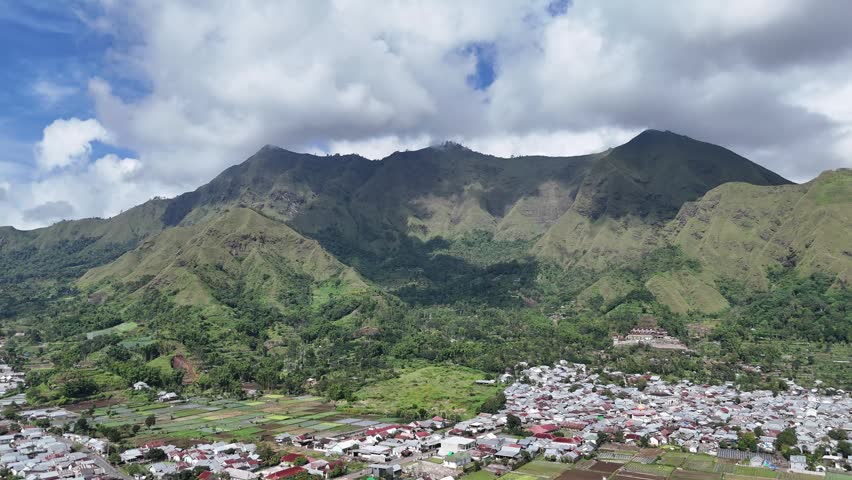 Aerial view of a stunning green mountain range towering over a vibrant town and agricultural fields, under a dynamic cloudy sky, showcasing a blend of nature and human settlement.