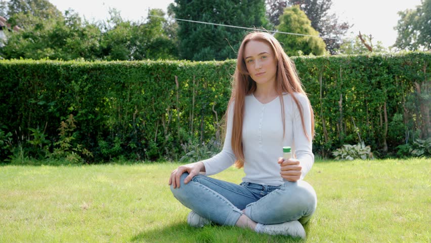 A girl sits on the grass and plays with a bubble wand. She blows bubbles and smiles. The sun shines bright as she enjoys her time outside in the garden.