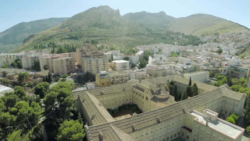 Aerial drone view of Catedral de Jaén in Jaén, Spain, glowing under bright summer sunlight with surrounding mountains and historic Renaissance architecture, cinematic 4K cityscape, clear blue sky, detailed stone facade, peaceful urban panorama.