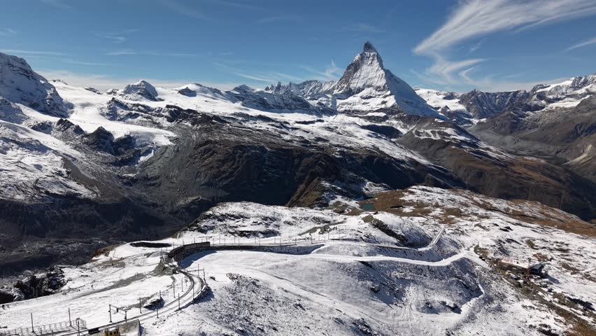 Aerial flyover over alongside the Gornergrat railway in Zermatt, Switzerland with a view of a train riding down the tracks and the Matterhorn on a snowy autumn day in the Swiss Alps