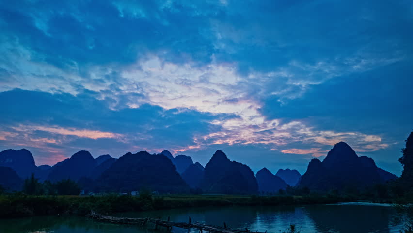 The calm stream reflects the fiery colors of the setting sun as a wooden bridge connects the fields, with lush greenery and majestic peaks completing this peaceful countryside scene.