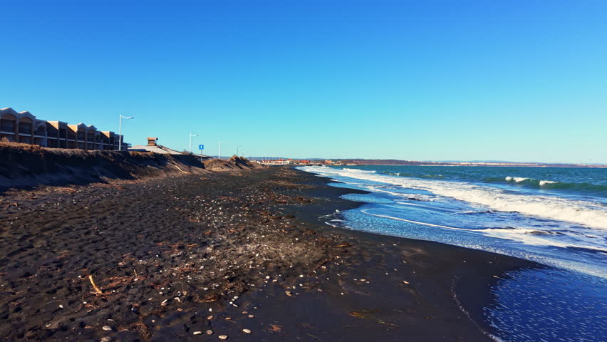 People stroll along the beach with black sand as the waves crash gently against the shore under a clear blue sky during the day.