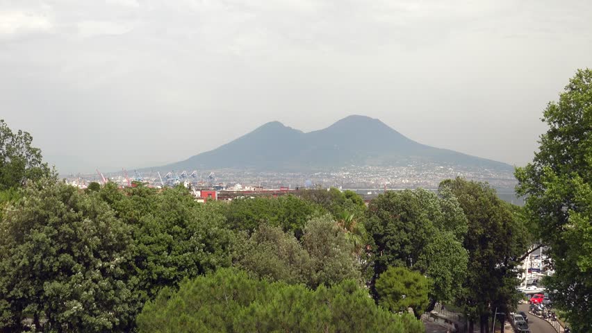 Zoom in view of Naples Italy reveals Mount Vesuvius rising beyond green urban landscape. greenery gives way to volcanic silhouette, italian skyline, landmark scenery, travel appeal. Mount Vesuvius is an iconic Campania stratovolcano near Naples, linking the skyline with volcanic geology, Mediterranean settlement, historic eruption memory, and Italian landmark scenery.