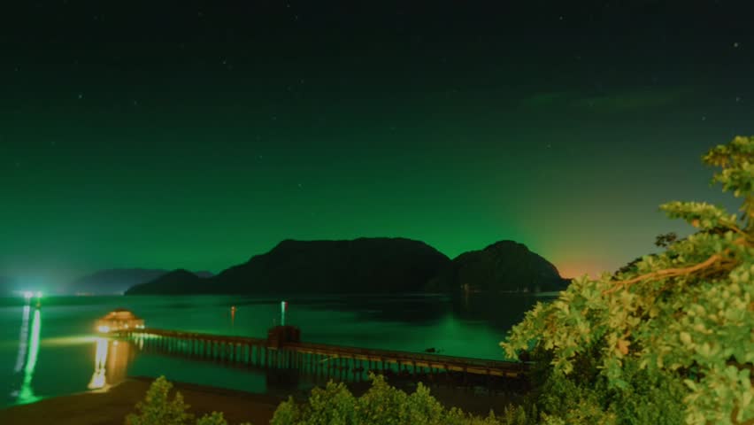 A stunning night view of the beach with a dramatic green light phenomenon in the sky, resembling the Aurora Borealis. In the foreground, there is a long wooden jetty jutting out into the calm sea.
