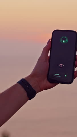 Vertical video. Woman Holding Smartphone Showing Satellite Search Interface during Ocean Sunset at Cliff Edge Demonstrating Starlink Satellite Communication Technology in Remote Travel Location.