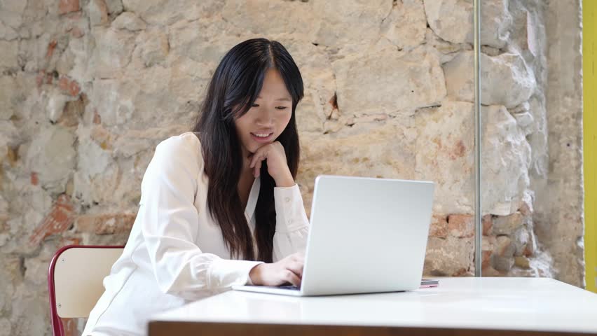 Young woman works calmly at laptop. Her expression shows focus and mild concentration. Stone wall background adds rustic charm. She sits at clean white table, fingers moving over keys