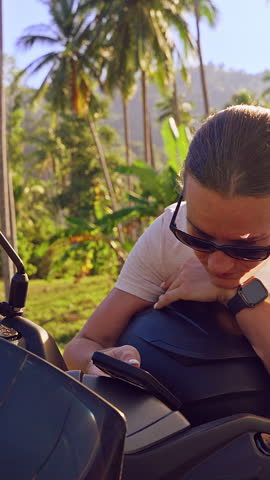 Vertical video. Young Woman Leaning on Scooter and Looking at Smartphone Screen on Tropical Road With Palm Trees and Hills in Background, Searching Route and Planning Travel Direction.