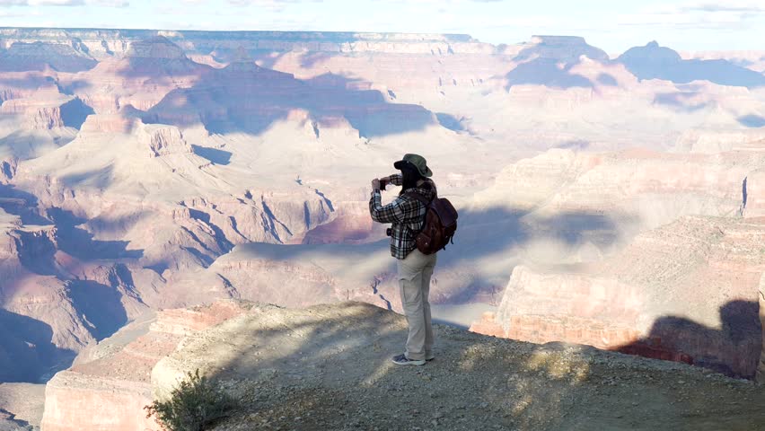 Young female traveler with a backpack standing on the edge of a cliff, taking pictures of the breathtaking panoramic view of the grand canyon national park with her smartphone