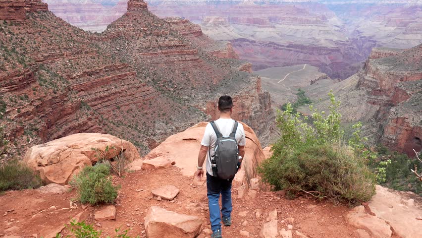 Young male backpacker walking to the edge of a cliff at grand canyon national park, raising his arms in a gesture of freedom and success while admiring the breathtaking panoramic view