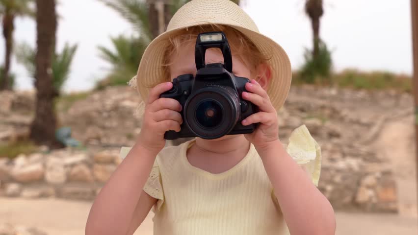 Close up of a little girl in a straw hat holding a professional DSLR camera in front of her face. Toddler pretending to take photos during a bright sunny day at an outdoor park with palm trees