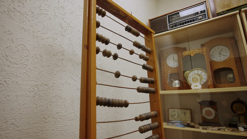 Vintage abacus stands in foreground of historical museum room. Background features glass case filled with antique clocks and retro radio