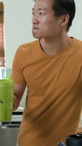Vertical video: Reacting to joke, man in orange tee with blender cup, high-fiving friend in kitchen. Candid, smoothie, green, casual, interior, sociable, vertical video