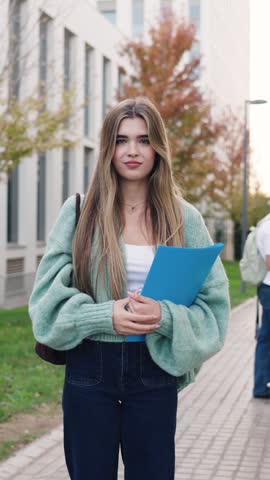Portrait of a young female student with long hair, smiling and looking at the camera. She is holding a blue folder in her hands. Vertical