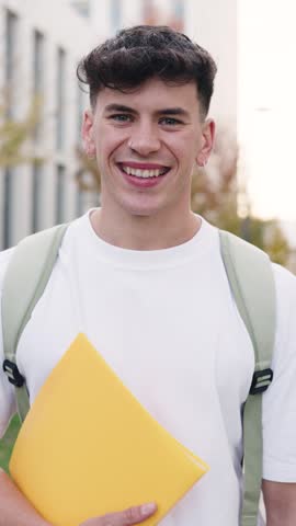 Confident young male student with a backpack holding a yellow folder. Handsome happy man looking at the camera while standing in the campus. Vertical