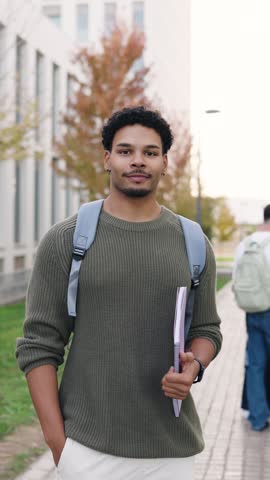Portrait of a confident African American male student with a backpack and notebook smiling at the camera on the college campus grounds. Vertical