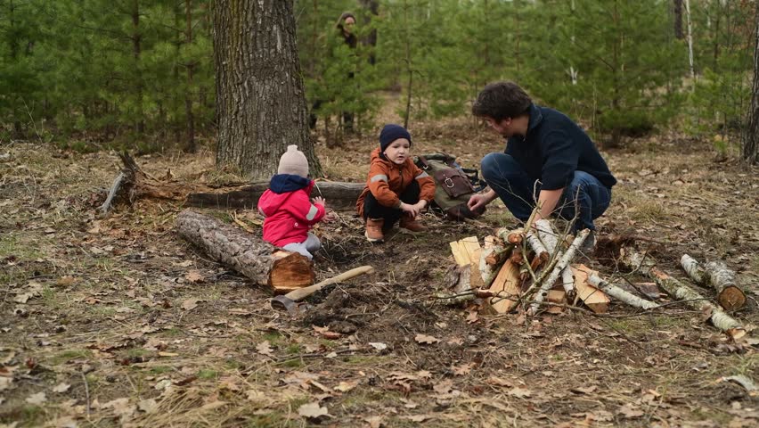 Father and children digging the ground in the forest to prepare a future campfire area. Firewood lies nearby, creating a warm outdoor family and camping scene in nature.