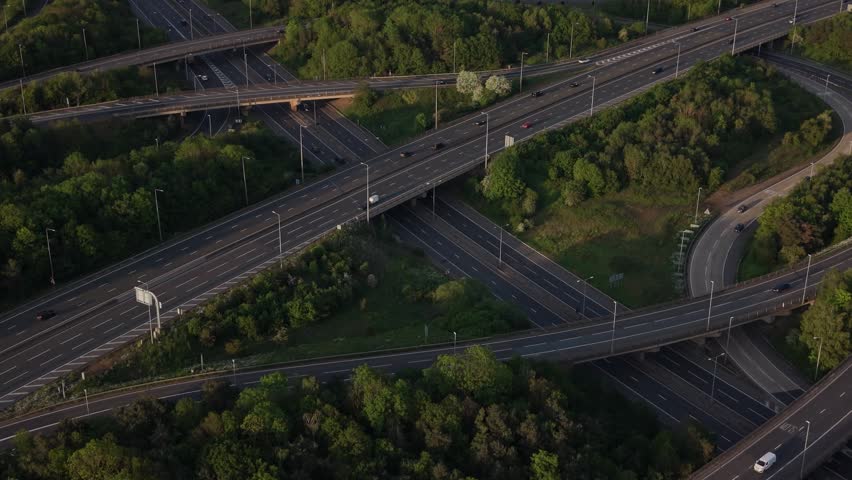 Overhead perspective of a complex multi-level highway interchange with vehicles moving through various roads and ramps. Lush green trees line the infrastructure.