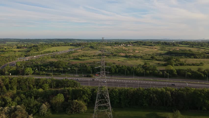 An aerial perspective shows cars moving on a busy highway through lush green countryside. A tall power line tower stands prominently amidst fields and distant trees under a bright daylight sky.