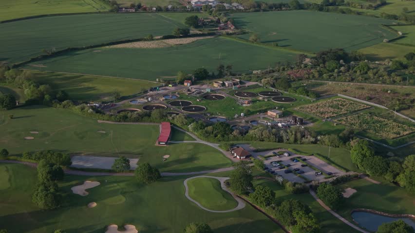 Panoramic view captures a vibrant green golf course with bunkers and ponds. Nearby, a complex of circular water treatment tanks stands amidst rolling fields.