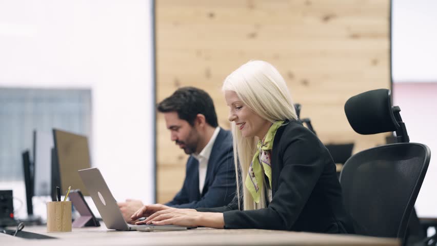 Two focused office workers sitting at a desk and typing on their laptops. Modern and professional environment for business collaboration and productivity