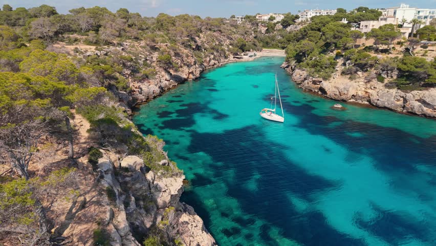 4K aerial: Turquoise water fills narrow rocky cove of Cala Pi, Mallorca, Spain. Pine trees cover steep cliffs in Cala Pi, Mallorca, Spain. Boats anchor in natural sea inlet of Cala Pi, Mallorca, Spain