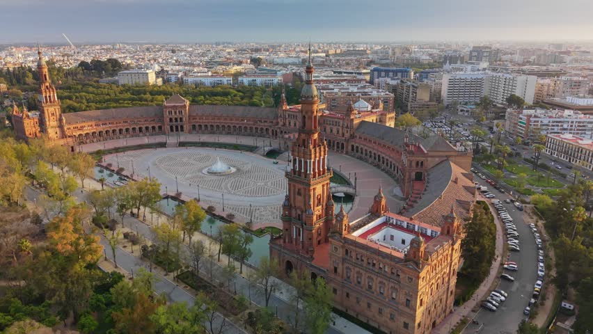 Central fountain flows in Plaza de Espana, Seville, Spain. Historic brick towers surround square in Seville, Spain in this aerial UHD. Water canal borders semicircular architecture in Seville, Spain