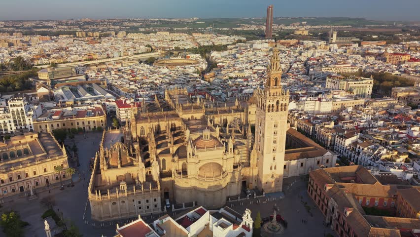 Giralda bell tower rising over Cathedral of Seville, Spain. Golden sunset hits Gothic roofs of cathedral in Seville, Spain. Dense cityscape surrounds historic cathedral in Seville, Spain. Aerial 4K