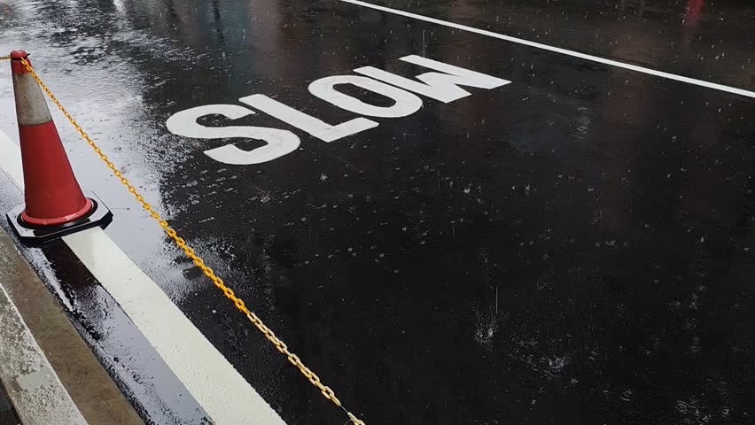 A rainy day showing raindrops hitting a dark, wet asphalt road. A large white "SLOW" traffic sign is painted on the ground next to a red traffic cone and yellow chain.