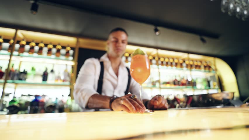 Charismatic professional barman serving a delicious orange cocktail to a customer. An expert mixologist working behind the counter at a bar