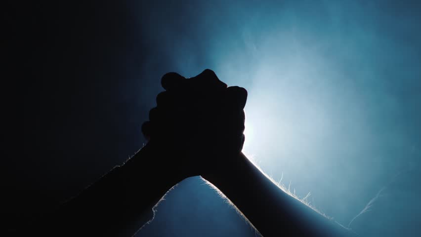 Close up silhouette of two people joining hands together in arm wrestling pose against bright blue glowing light. Cinematic footage depicting unity support partnership connection and strength.
