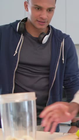 Vertical video: Looking down man in hoodie reaching blender and fruit, making drink at home. Kitchen, modern, preparation, banana, apple, lighting, vertical video