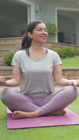 Vertical video: Settling woman in gray top, leggings doing neck rolls on pink mat patio, calming. Yoga, meditation, lotus, barefoot, lavender, garden, vertical video